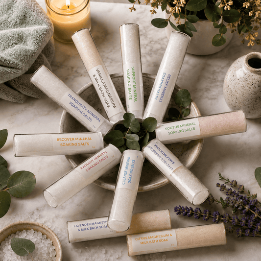 Ten glass tubes filled with Elixir Mind Body Botanicals 10 bath soak scents 7 mineral salt soaks, and 3 magnesium baths laid out over a table with towel plants and bowl of salts