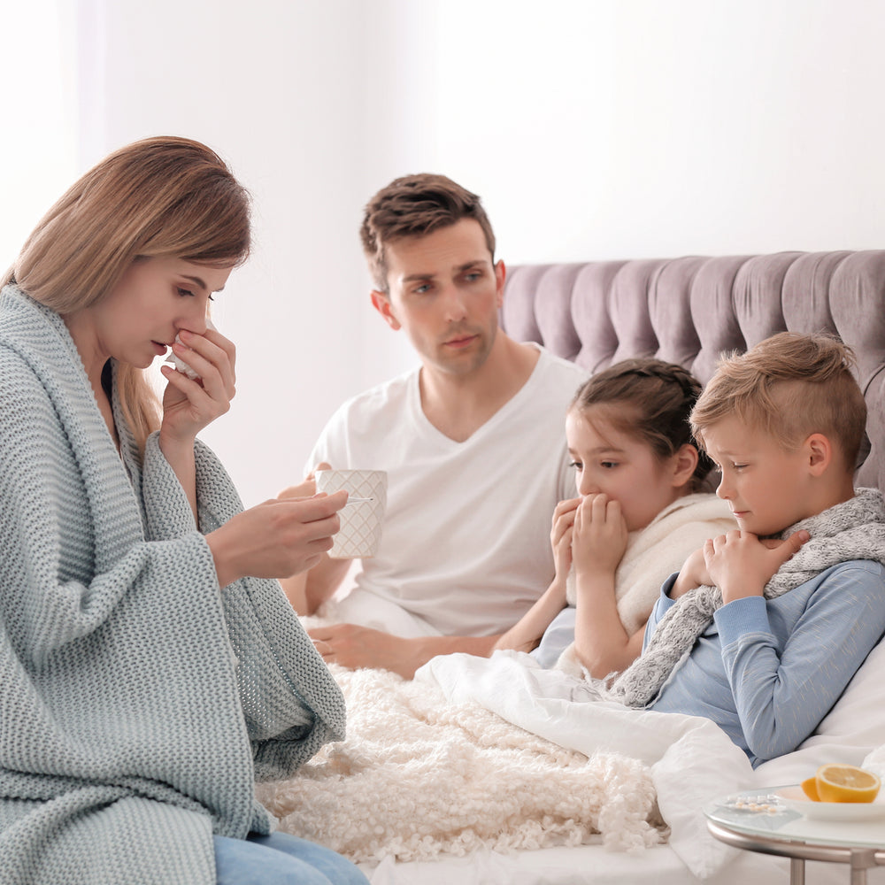 Family of four sitting in bed together, with a woman holding a thermometer and tissue, a man holding a mug of tea, and two children wrapped in blankets.