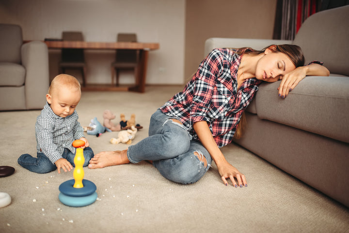 A young, exhausted motheris  on the floor with her toddler, and has fallen asleep while he plays with his toys