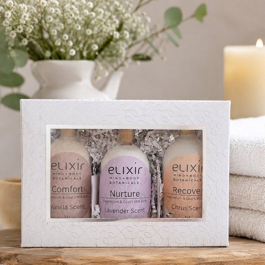 Elixir Mind Body Botanicals Magnesium & Goat's Milk Bath glass-bottle gift set in a white box on a wooden shelf, with folded white towels and flowers in a vase.