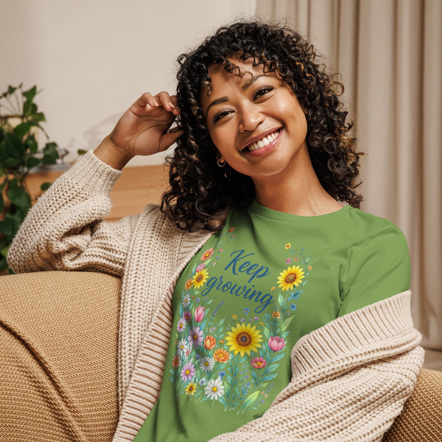 Front view of a smiling woman sitting on a chair wearing a tan sweater over her relaxed light green short-sleeved t-shirt with a colorful, floral design that says "Keep Growing."