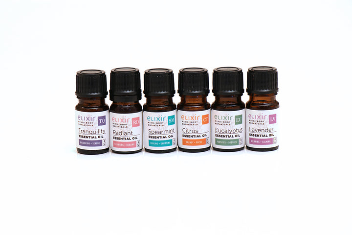 A series of six small bottles of Elixir Mind Body Botanicals Essential Oils, each labeled with different names such as Citrus, Tranquility, Radiant, and others. The bottles are arranged in a line against a white background.