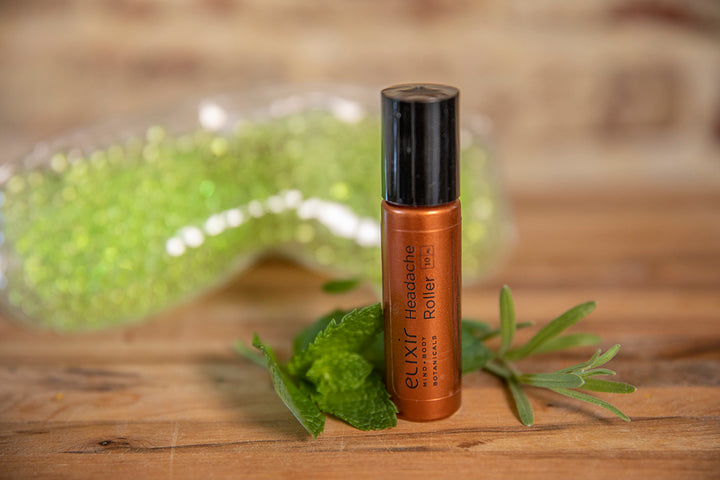 Elixir Mind Body Botanicals Headache Roller on a butcher block counter with mint and lavender leaves