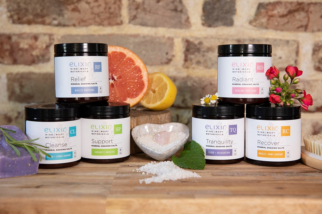 Elixir Mind Body Botanicals Mineral Soaking Salts in each scent: Relief, Cleanse, Support, Radiant, Tranquility, and Recover, grouped and stacked together with sliced citrus, mint leaves, dry brush, geranium buds, lavender spring, and heart dish with mineral salts and crystal on a butcherblock countertop and a brick background