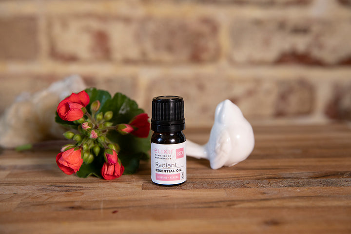 Elixir Mind Body Botanicals Radiant Essential oil on a butcher block countertop with geranium bloom and a small white stone bird