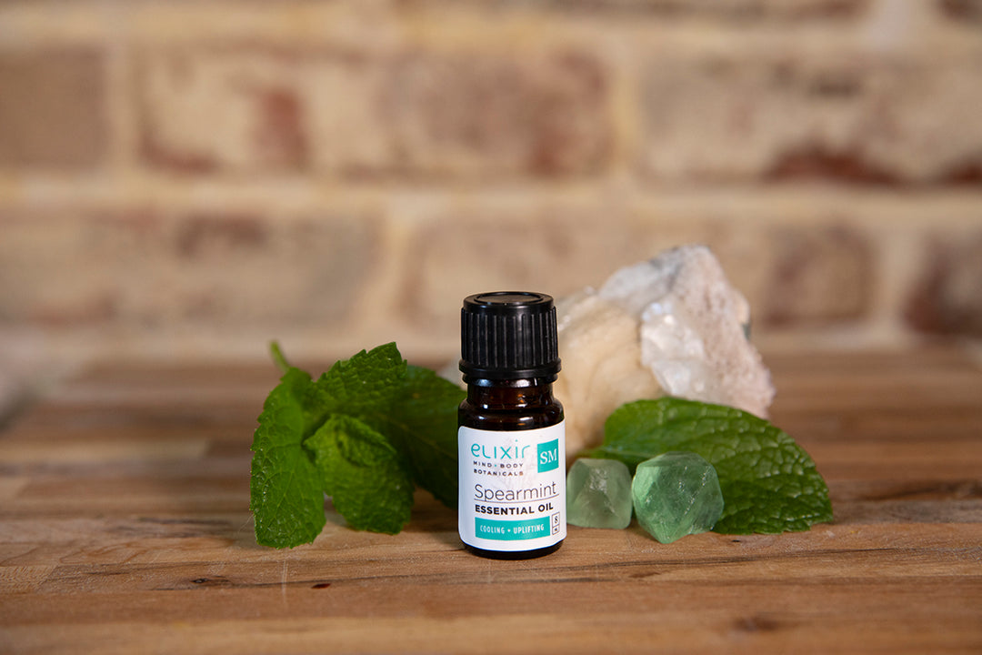 Elixir Mind Body Botanicals Spearmint Essential Oil on a butcher block counter with mint leaves and green crystals