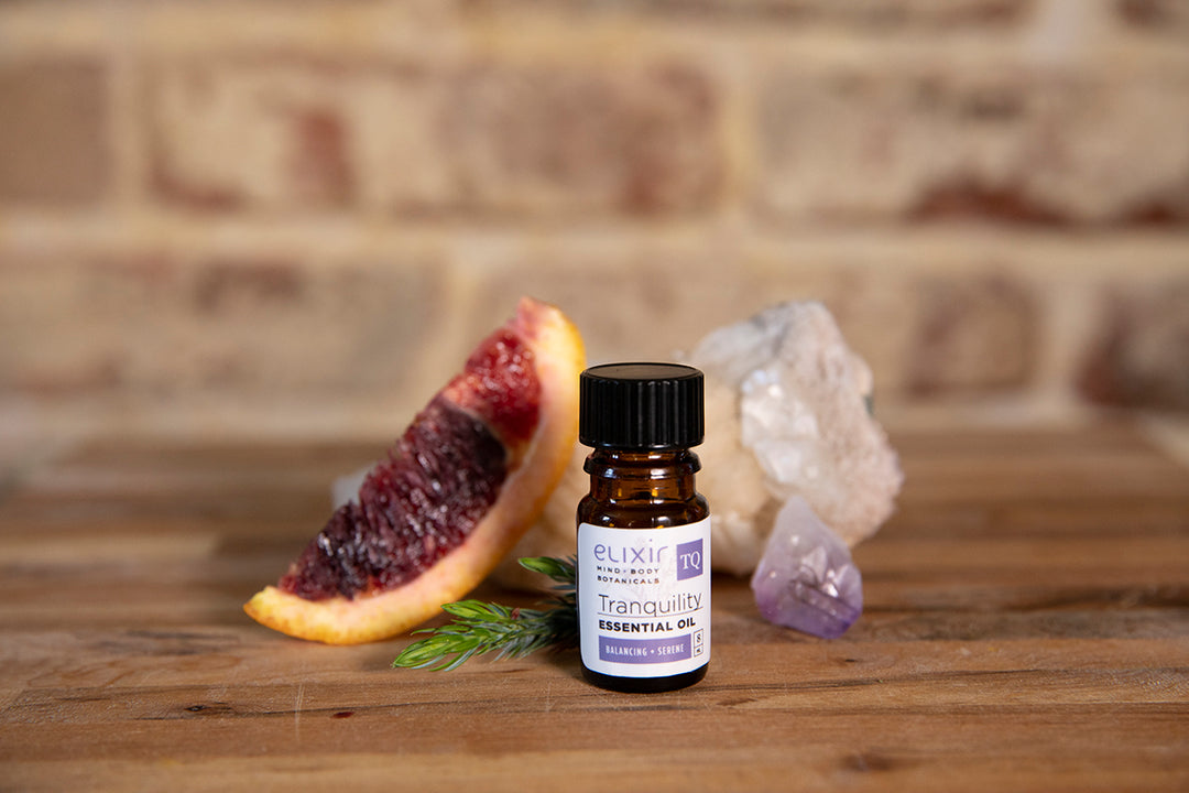 Elixir Mind Body Botanicals Tranquility Essential Oil on a butcherblock counter with blood orange slice, juniper sprig, and crystals