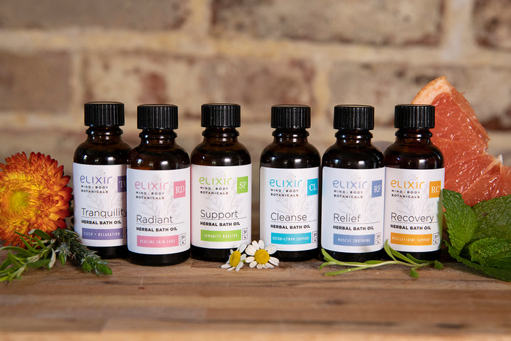 Group of 6 Elixir Mind Body Botanicals Herbal Bath Oils: Tranquility, Radiant, Support, Cleanse, Relief, and Recovery on a butherblock countertop with herbs, flowers, and citrus slices