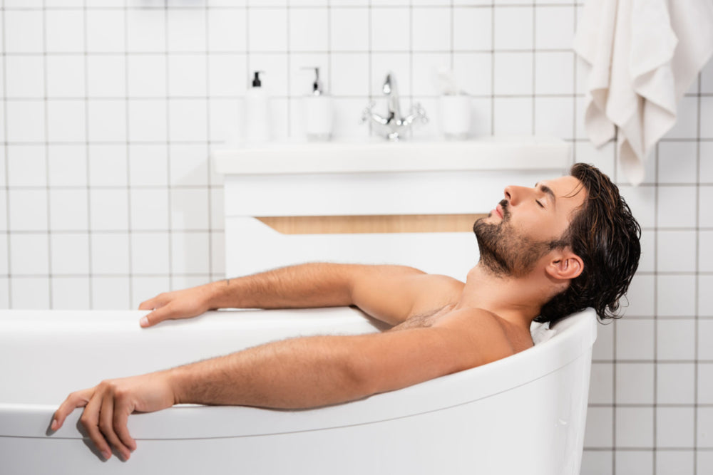 A man with a beard is soaking in a tub in a white tiled bathroom using Elixir Mind Body Botanicals Mineral Soaking salts with maganesium