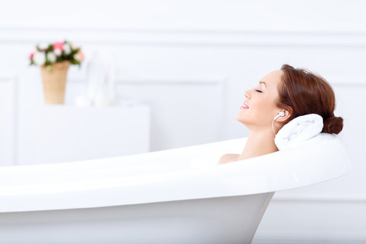Redheaded woman relaxing in a white bathtub with a rolled towel under her neck and earbuds in, listening to music