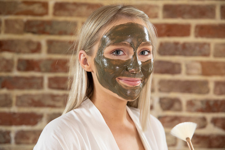 Blonde woman in a robe using Elixir Mind Body Botanicals Mineral Mud Mask on her face 